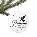 Front view of White ceramic Christmas ornament with graphic displaying stars, an angel, and text that says Believe. Hanging on a branch.