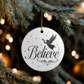 Front view of White ceramic Christmas ornament with graphic displaying stars, an angel, and text that says Believe. Hanging in a tree