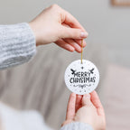 Front view of White ceramic Christmas ornament with graphic displaying stars, angels, and text that says Merry Christmas. Model holding ornament with both hands