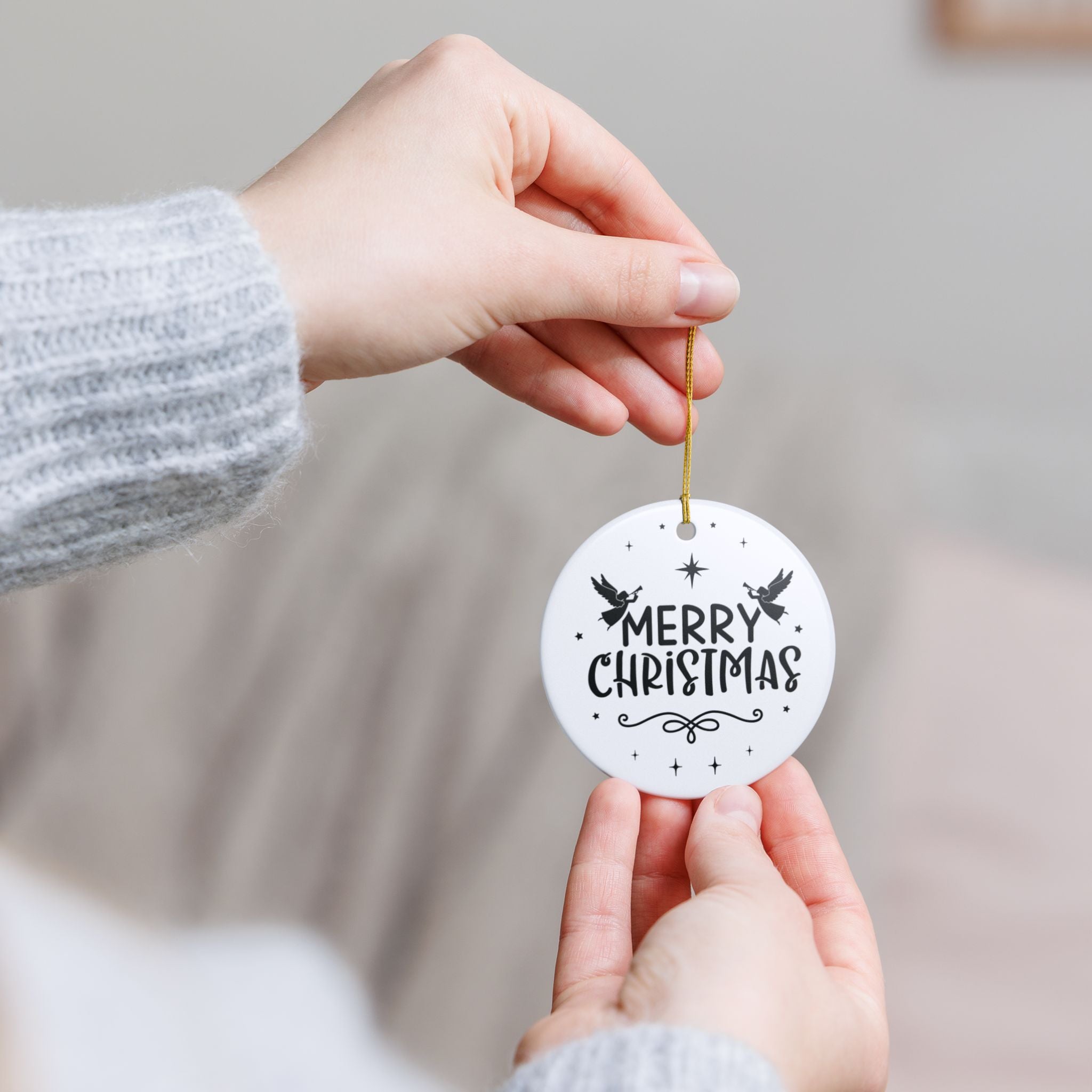 Front view of White ceramic Christmas ornament with graphic displaying stars, angels, and text that says Merry Christmas. Model holding ornament with both hands