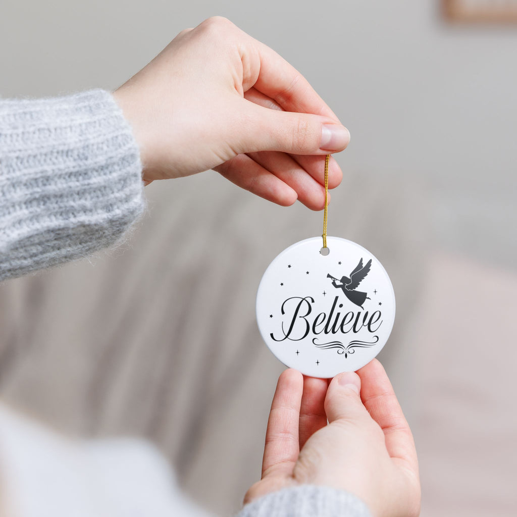 Front view of White ceramic Christmas ornament with graphic displaying stars, an angel, and text that says Believe. Model holding ornament with both hands.