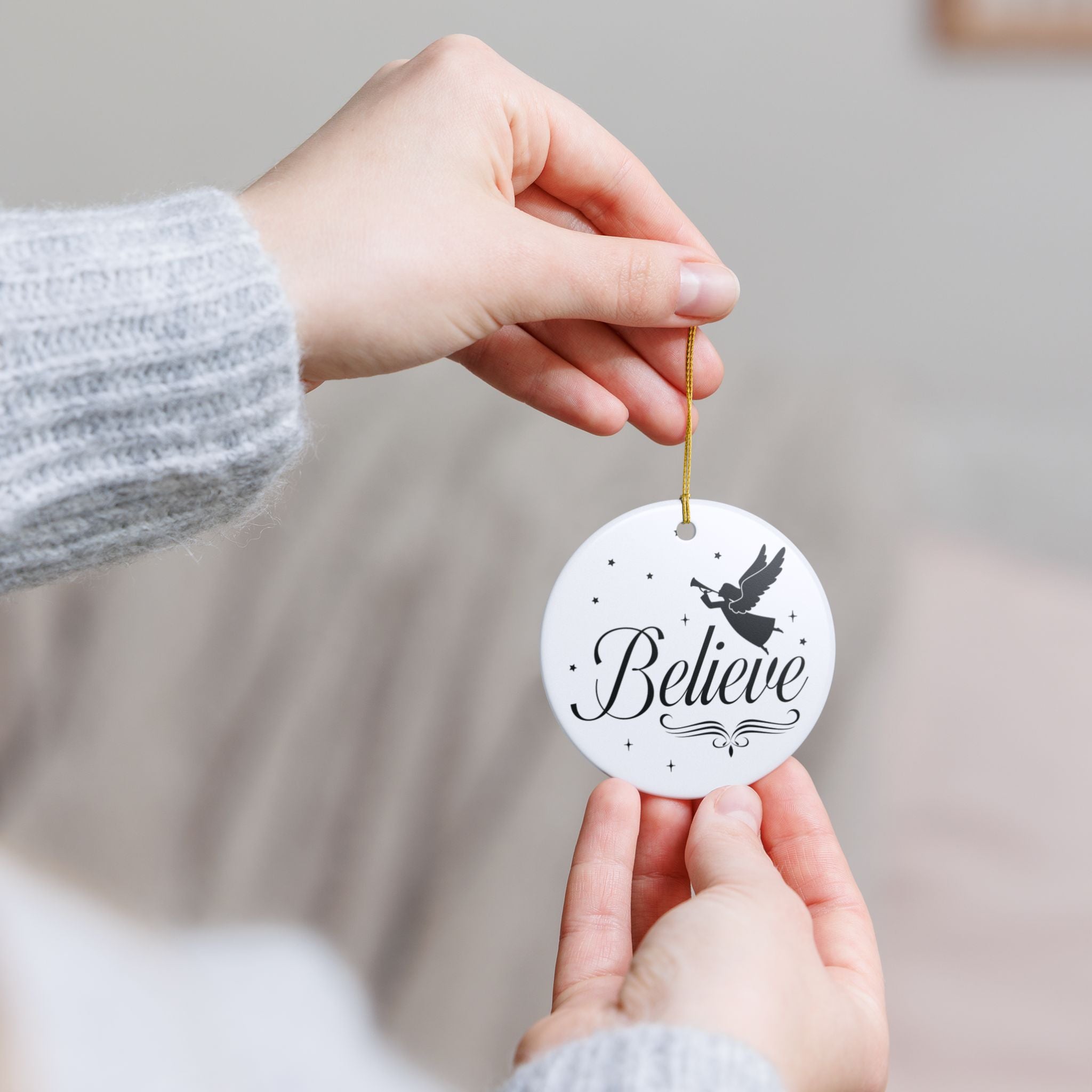 Front view of White ceramic Christmas ornament with graphic displaying stars, an angel, and text that says Believe. Model holding ornament with both hands.