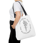 Front view of large White tote bag with black handles, large black floral graphic and Luke 12:27 Bible verse, shown on model's shoulder