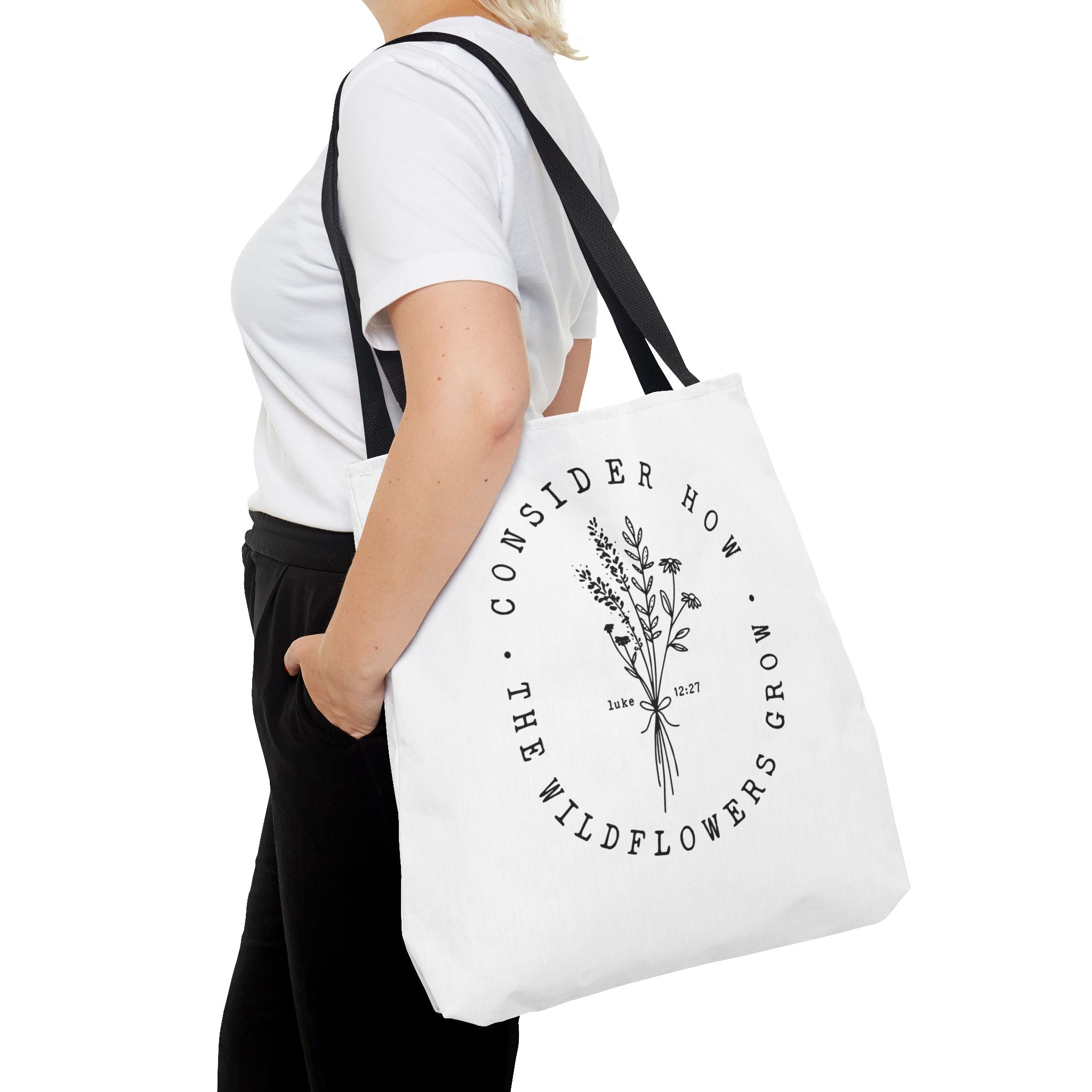 Front view of large White tote bag with black handles, large black floral graphic and Luke 12:27 Bible verse, shown on model's shoulder