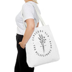 Front view of large white tote bag with white handles, large black floral graphic and Luke 12:27 Bible verse, shown on model's shoulder
