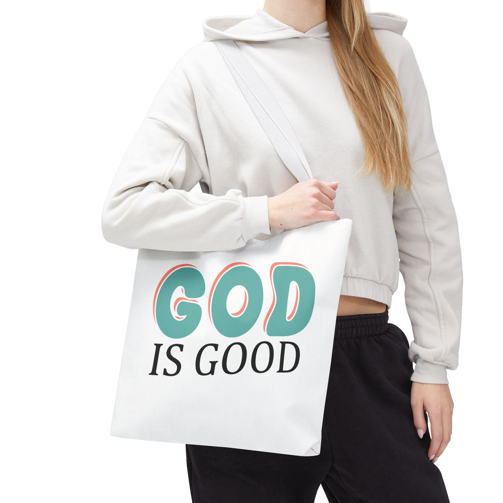 Front view of medium White tote bag with white handles, colorful graphic that says God is Good, displayed over model’s shoulder
