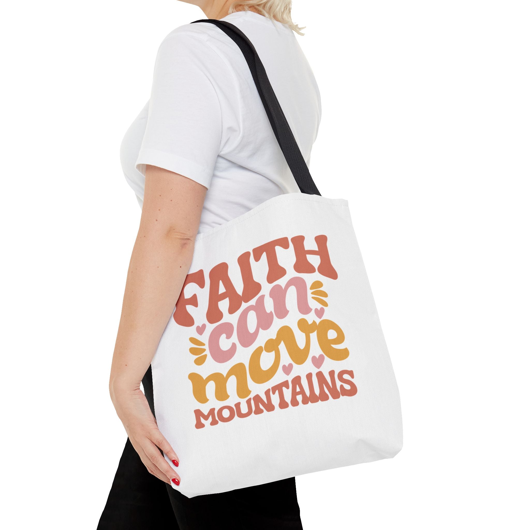 Front view of medium White tote bag with black handles, colorful graphic that says Faith Can Move Mountains, displayed over model’s shoulder