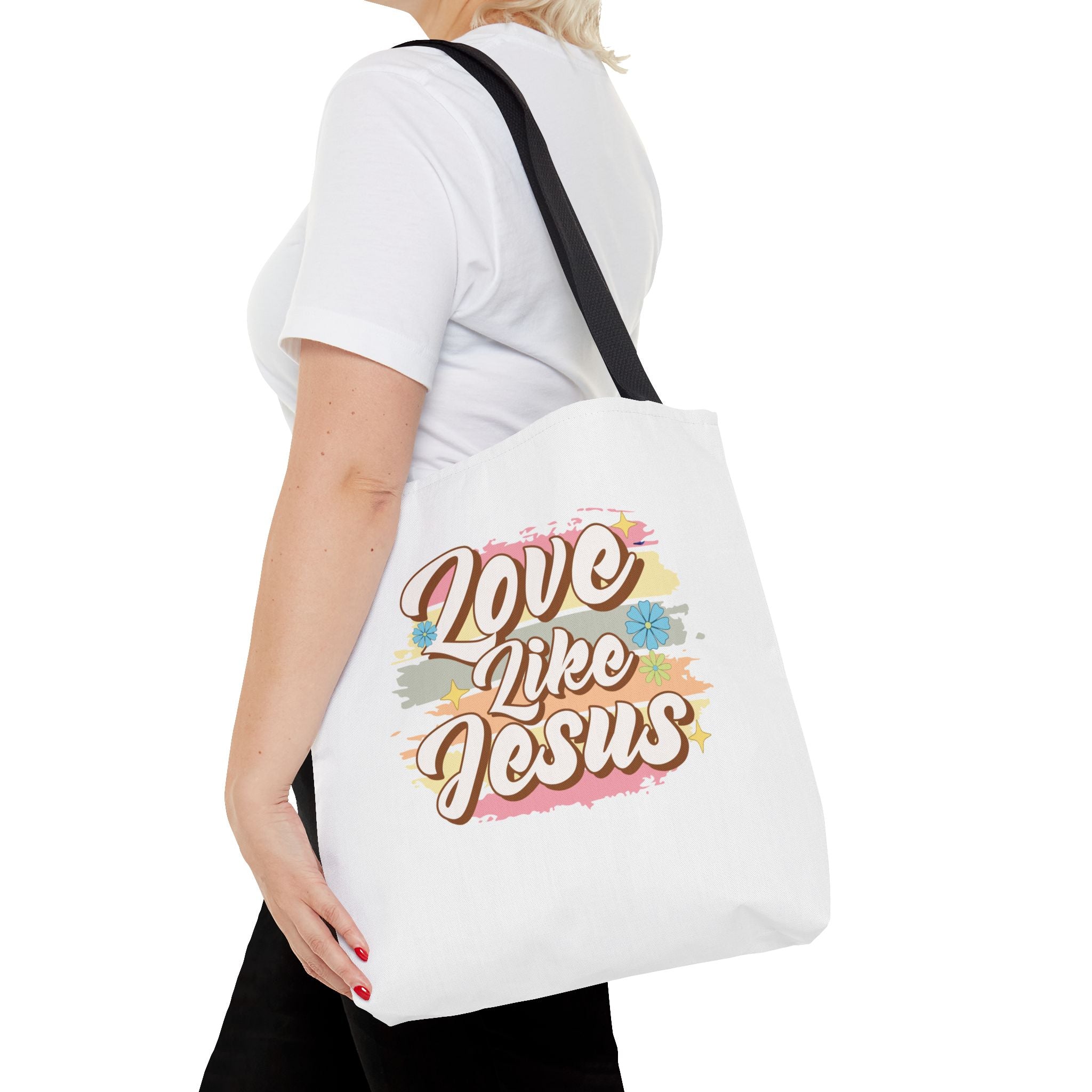Front view of medium White tote bag with black handles, colorful graphic with text Love Like Jesus, displayed over model’s shoulder