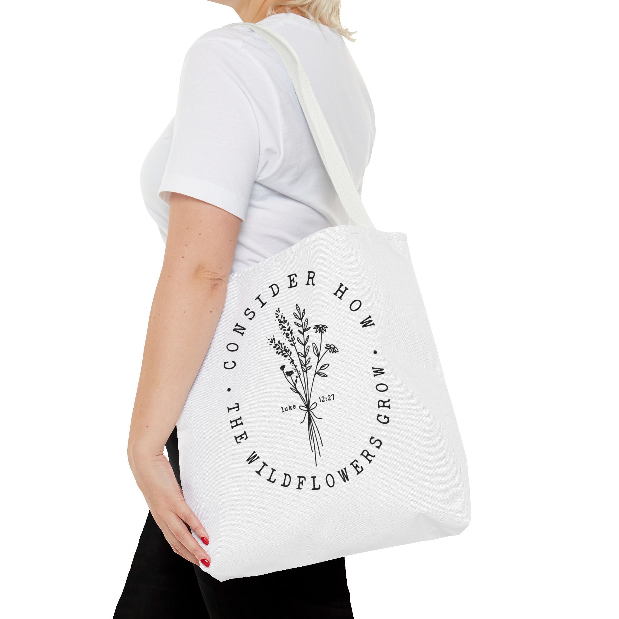 Front view of medium White tote bag with White handles, large black floral graphic and Luke 12:27 Bible verse, shown on model's shoulder