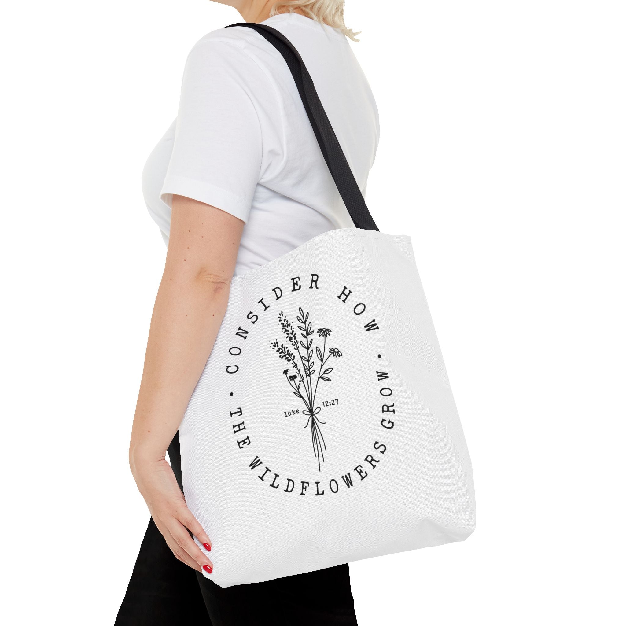 Front view of medium White tote bag with black handles, large black floral graphic and Luke 12:27 Bible verse, shown on model's shoulder