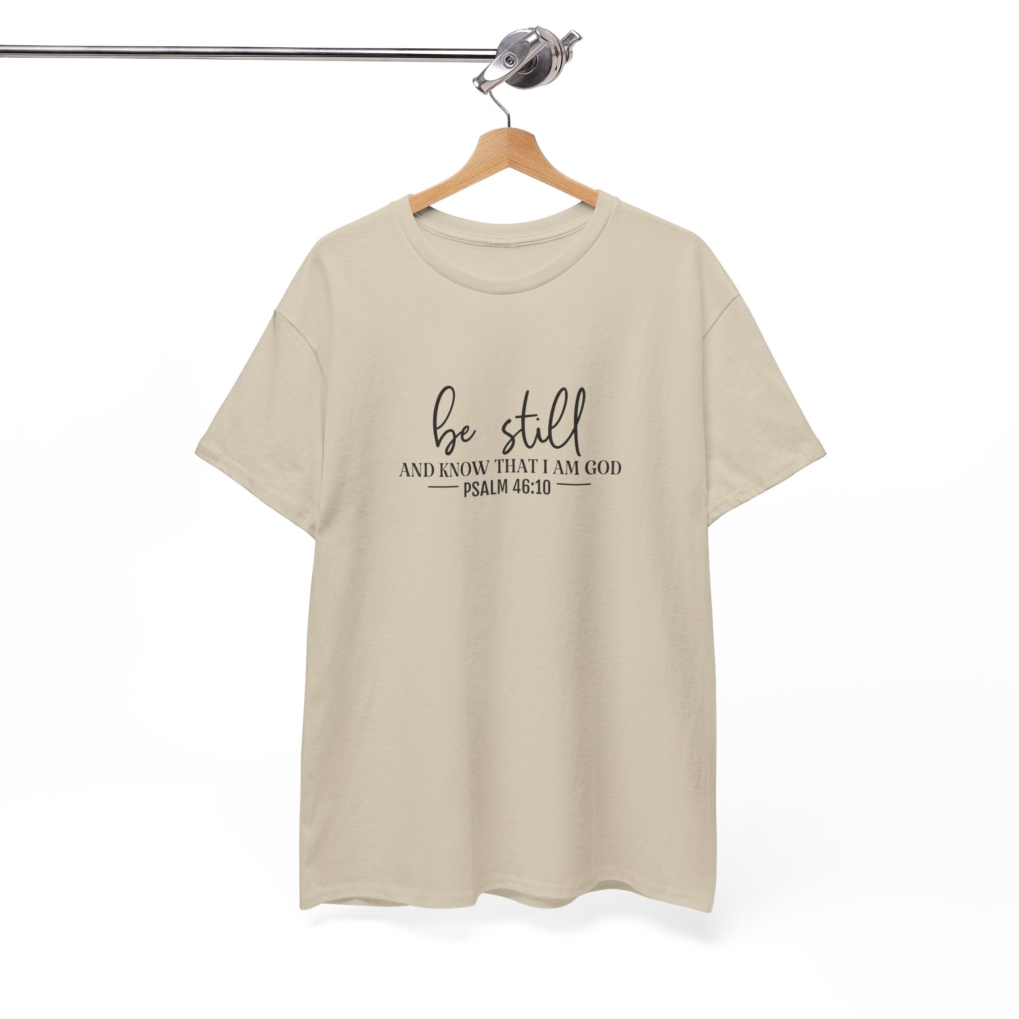 Front view of Sand t-shirt with black text Be Still And Know That I Am God Psalm 46:10 centered on chest. Shirt is displayed on a hanger.