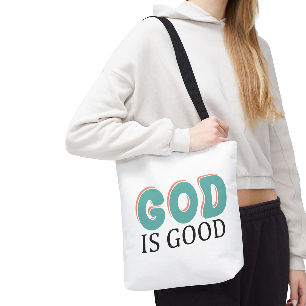 Front view of small White tote bag with black handles, colorful graphic that says God is Good, displayed over model’s shoulder