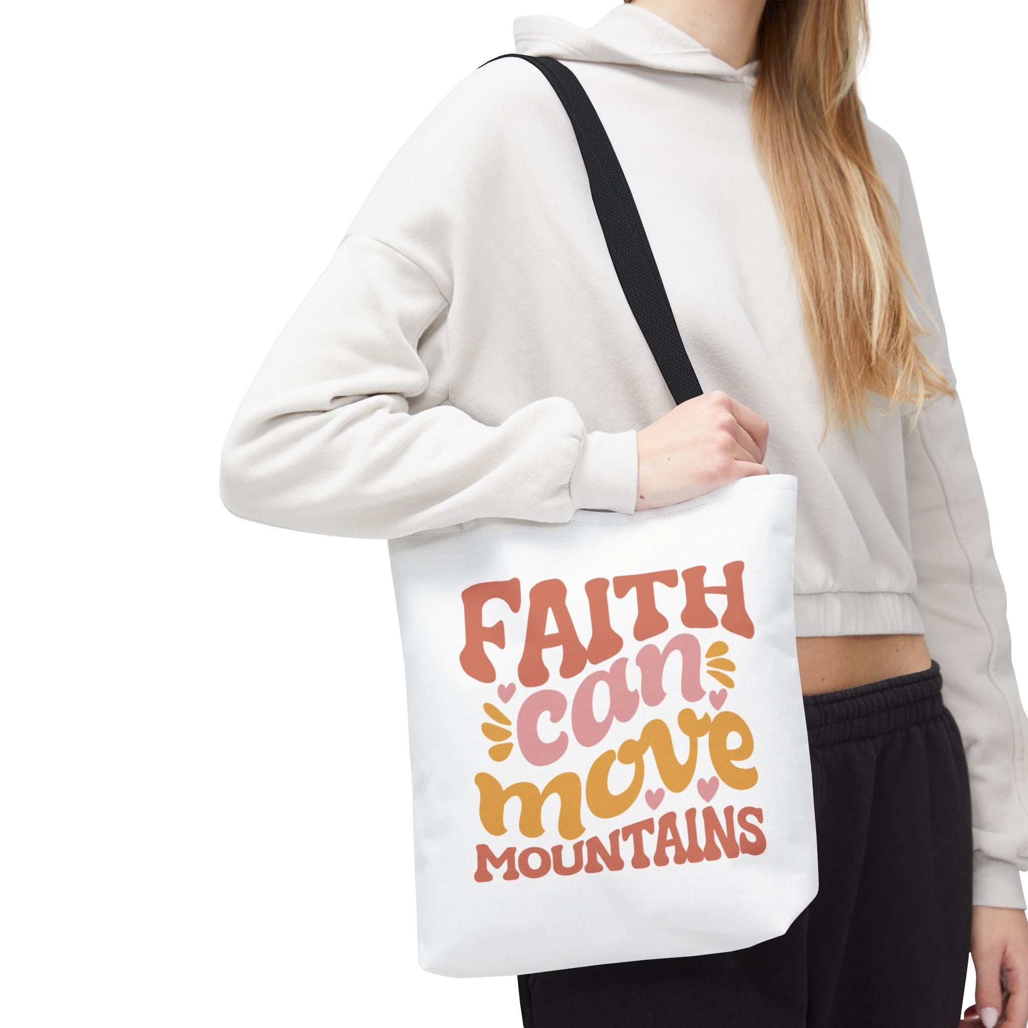 Front view of small White tote bag with black handles, colorful graphic that says Faith Can Move Mountains, displayed over model’s shoulder
