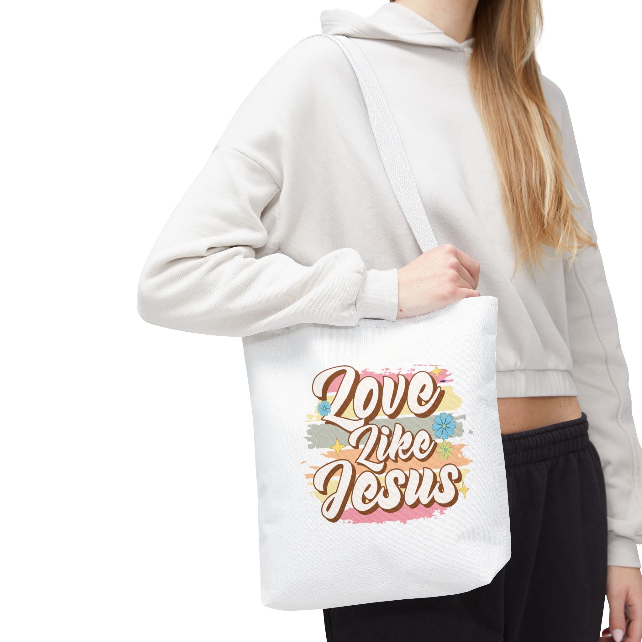 Front view of small White tote bag with white handles, colorful graphic with text Love Like Jesus, displayed over model’s shoulder