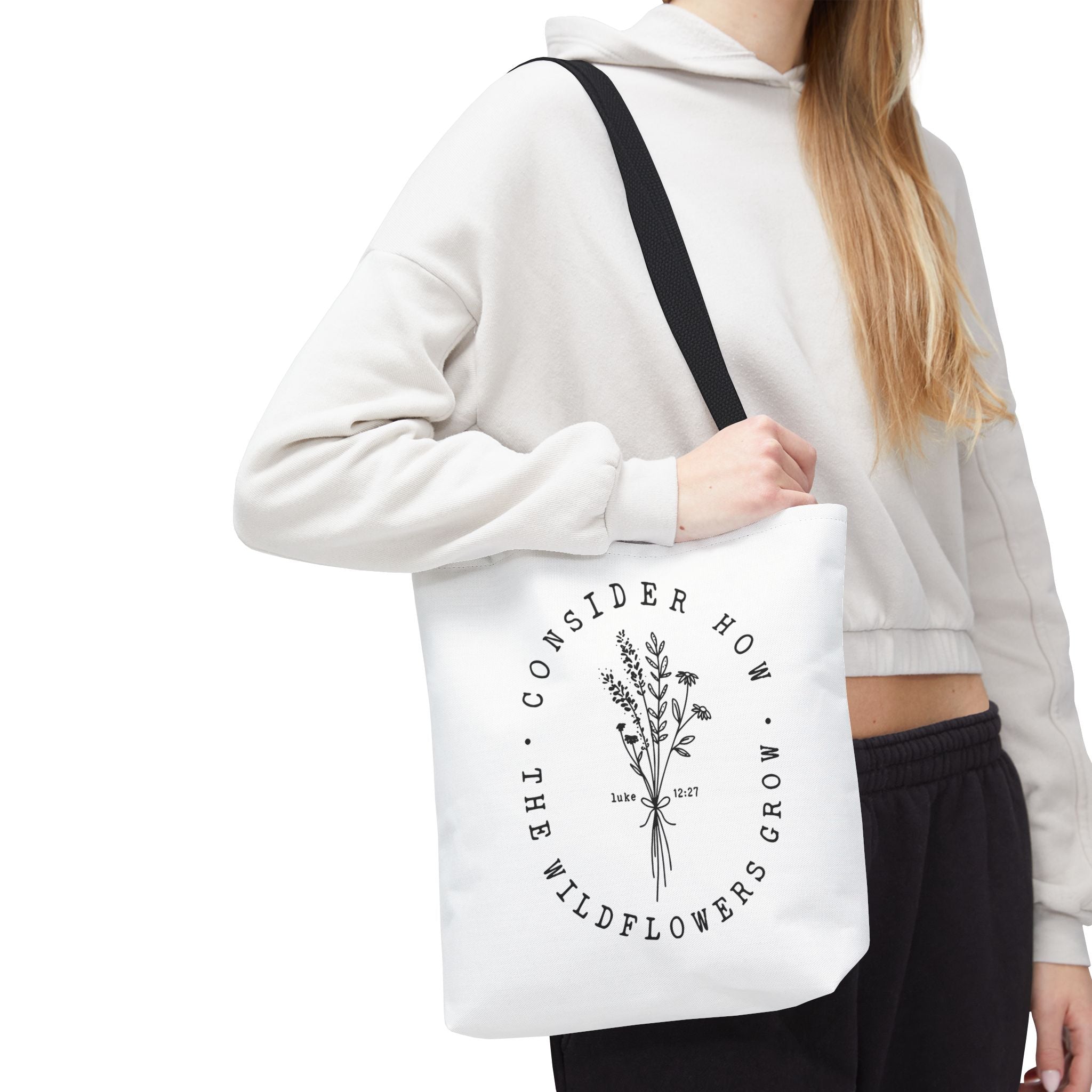 Front view of small White tote bag with black handles, large black floral graphic and Luke 12:27 Bible verse, shown on model's shoulder