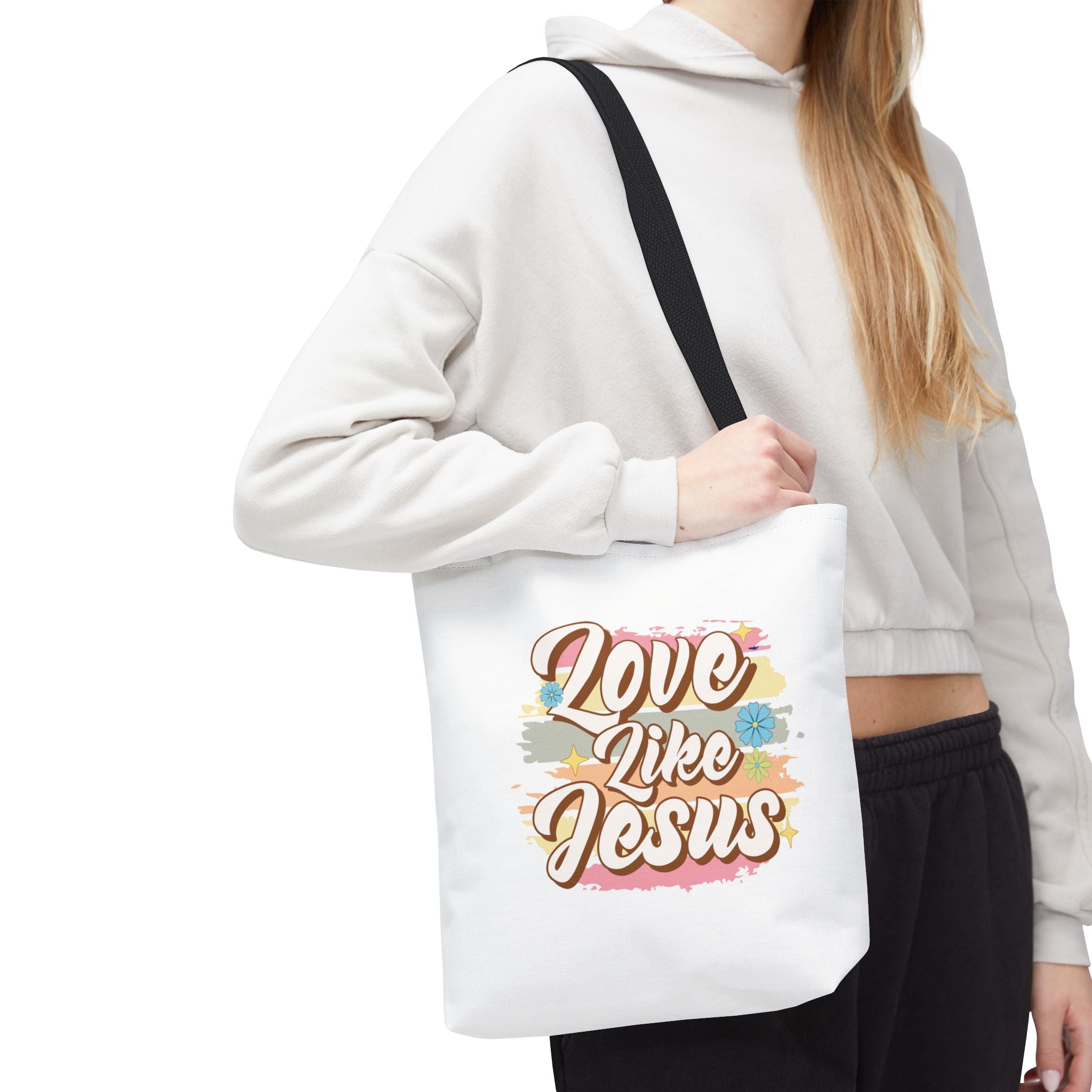 Front view of small White tote bag with black handles, colorful graphic with text Love Like Jesus, displayed over model’s shoulder