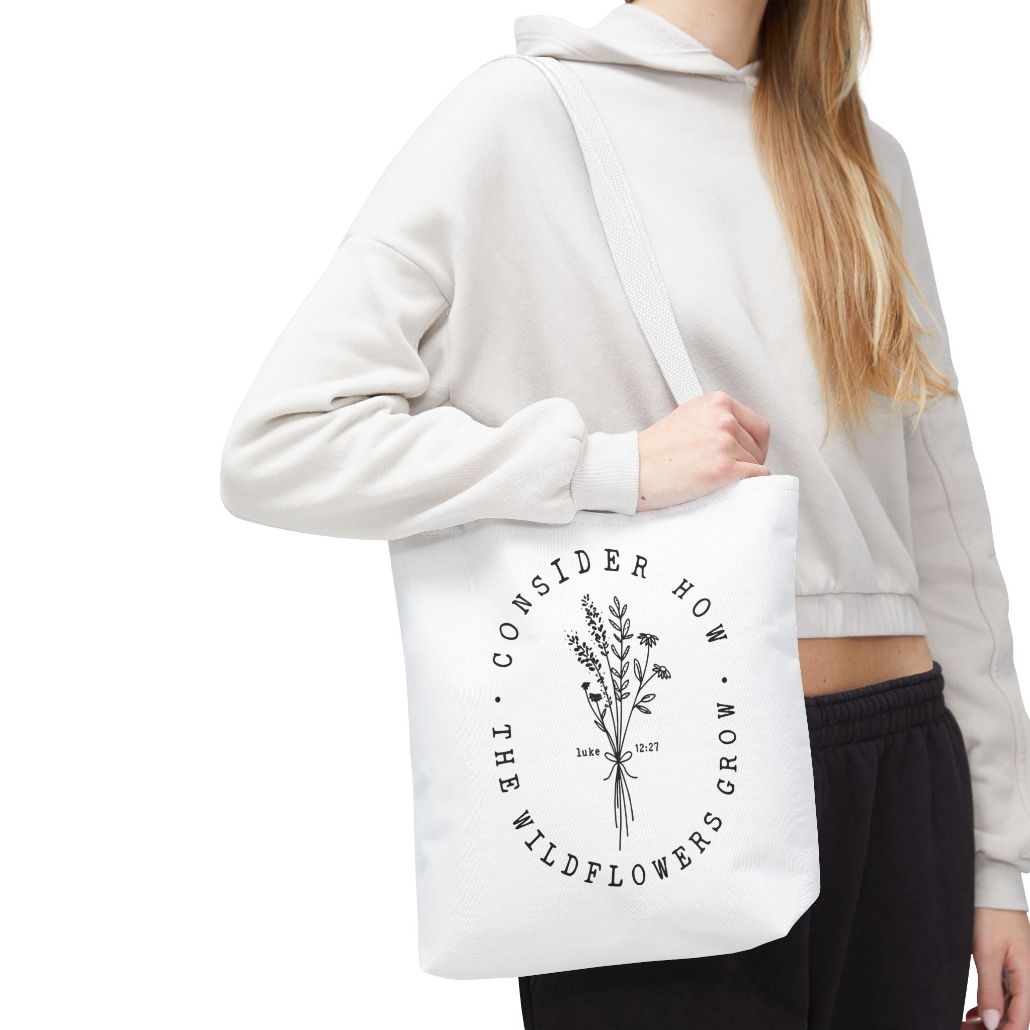 Front view of small White tote bag with white handles, large black floral graphic and Luke 12:27 Bible verse, shown on model's shoulder