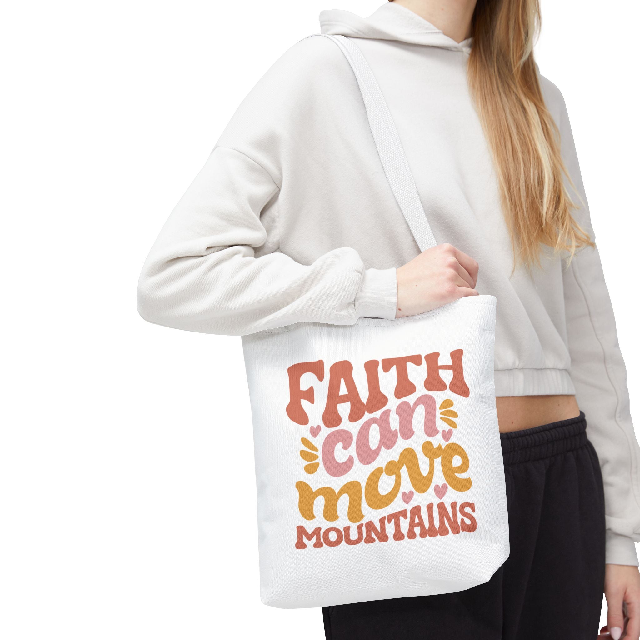 Front view of small White tote bag with white handles, colorful graphic that says Faith Can Move Mountains, displayed over model’s shoulder
